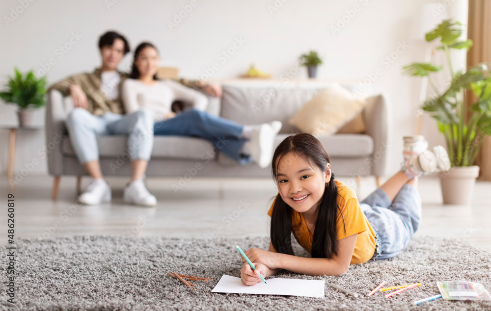 Happy teenager daughter lies on floor and draws, young asian lady and guy resting on couch in living room